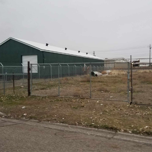 Green warehouse with a chain-link fence on a vacant lot with a cloudy sky.