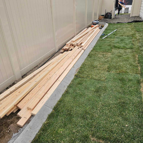 Wooden planks stacked next to a light-colored fence and green lawn. A person stands in the background.