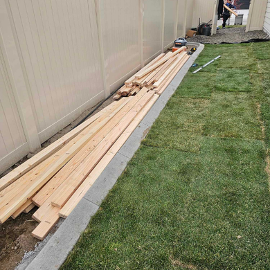 Lumber stacked along a tan fence next to a strip of grass and concrete. A person is visible in the background.