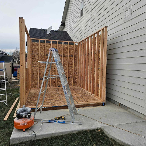 Shed under construction next to a house. Framing, air compressor, ladder, and tools are visible.
