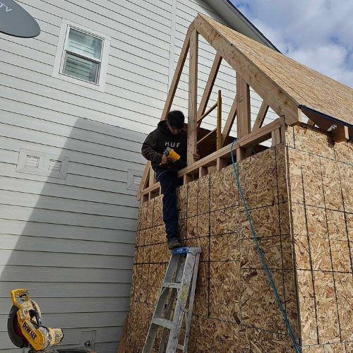A person using a nail gun on a wood structure attached to a white building. The person is standing on a ladder.