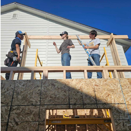 Three people building an exterior wall on a house. One uses a level, one checks measurements, and one has tools.