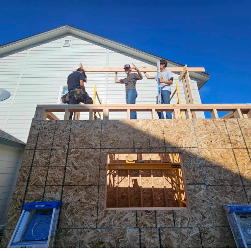 Three people building a wooden structure attached to a house; blue sky above.