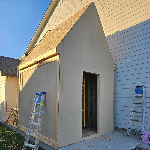 A shed under construction, built next to a house. Two ladders are present.