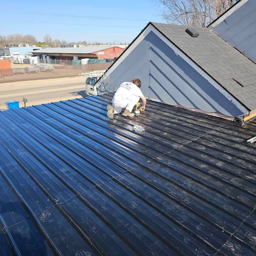 Person working on a black metal roof on a sunny day. Gray house in the background.