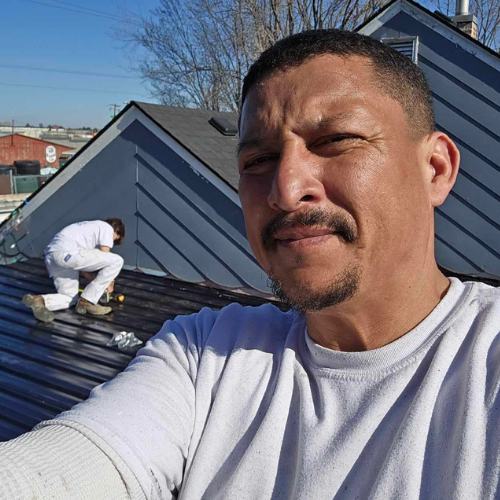 Man taking selfie on roof with another person painting siding. Blue sky, gray siding.