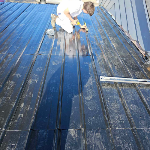 Person applies sealant to a black metal roof on a sunny day.
