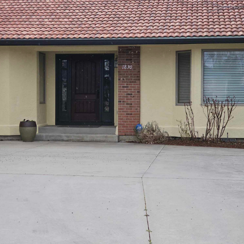 Beige house with red tile roof and dark front door, with concrete driveway.