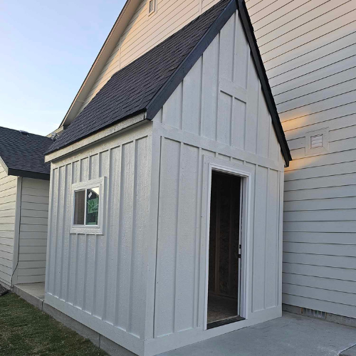 White-painted outdoor shed with a black roof, window, and open doorway, attached to a house with siding.