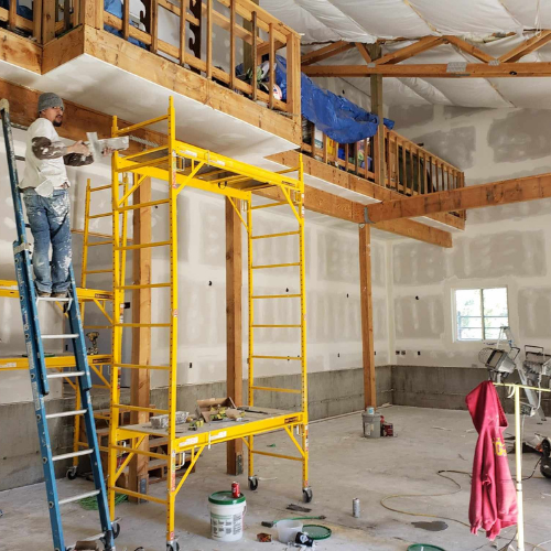 Man on ladder painting drywall in a large room with a yellow scaffold.