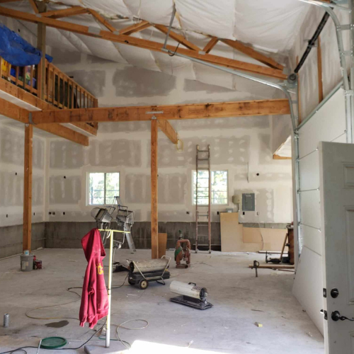 Interior of a large unfinished building with exposed wooden beams, drywall, and a partially open garage door.