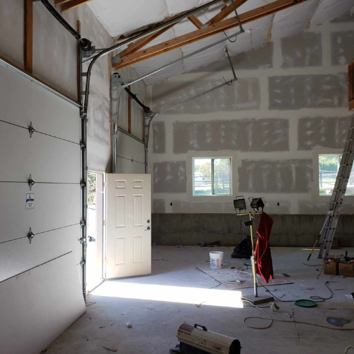 Interior of a partially constructed garage with a garage door open and a single interior door. Drywall is being installed.