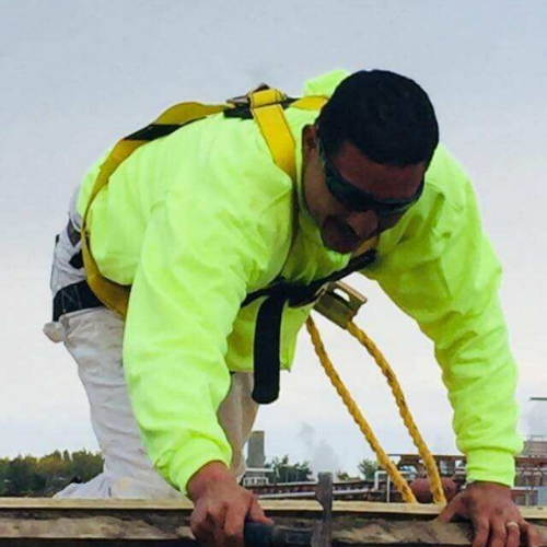 Construction worker in safety harness on a rooftop, wearing yellow shirt and sunglasses.