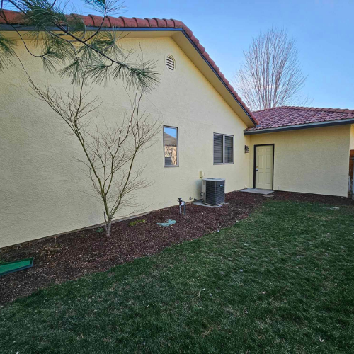 Yellow stucco house with red tile roof, small tree, grass, and storage shed.