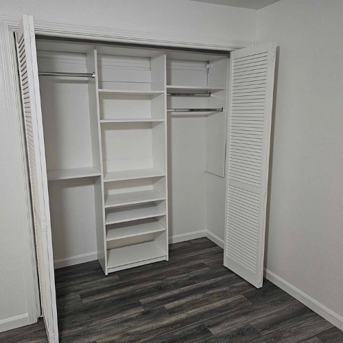 Empty white closet with built-in shelving and two sections for hanging clothes, open door on dark wood-look floor.