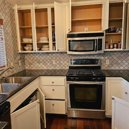 Kitchen with open cabinets, stovetop, microwave, and sink. White cabinets, stainless steel appliances, and dark countertops.