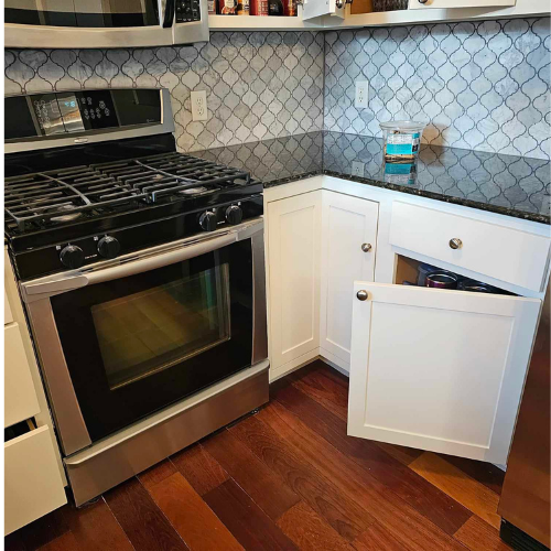 Kitchen corner with oven, white cabinets, and one door ajar. Dark countertops and patterned backsplash.