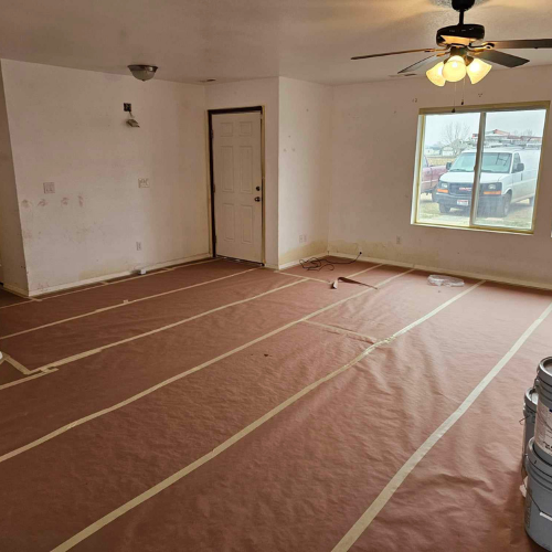 Room being renovated, with protective paper on the floor, white walls, and a window overlooking vehicles.