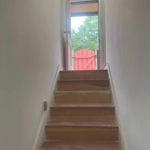 Wooden stairs lead to a doorway with a red gate. Exterior view with green trees.