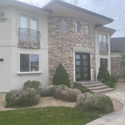 Two-story house with stone facade, dark doors, and small balconies, landscaped front yard with shrubs.