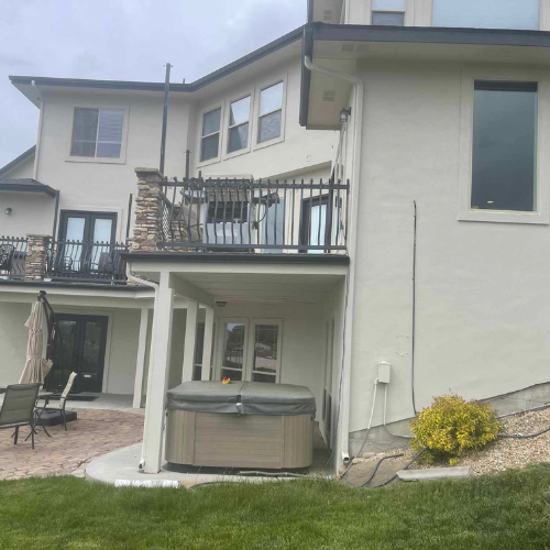 Beige house exterior with a hot tub under a second-story balcony; green grass in the foreground.