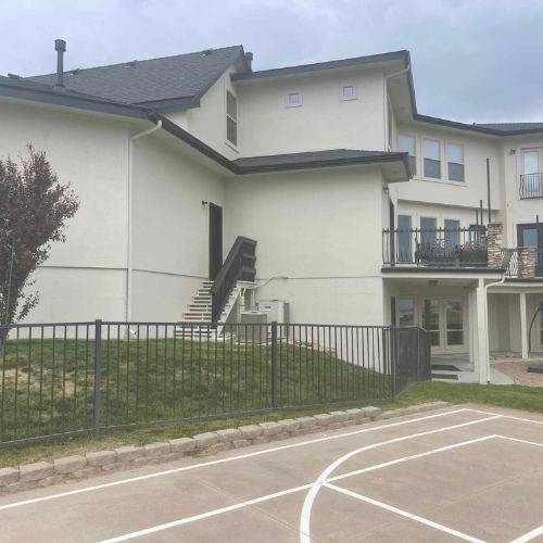 Two-story stucco building with dark roof, black metal fence, and outdoor basketball court. Stairs lead to a door.
