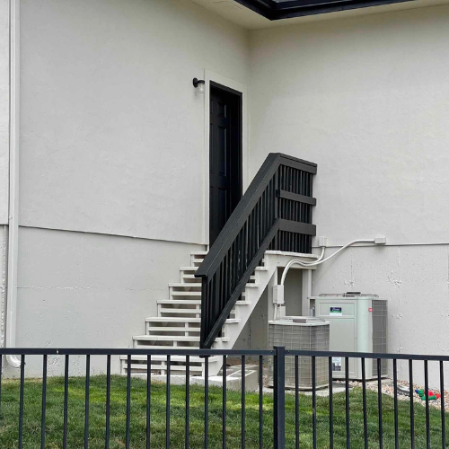 Exterior stairs leading up to a black door on a white building, with a black handrail and AC unit below.