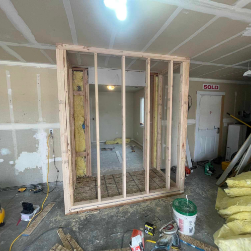 Framed wooden wall under construction in a garage, leading into a room with drywall and insulation visible.