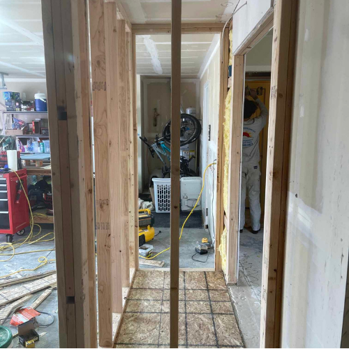 Hallway under construction with exposed wooden studs. Person working in the background. Tools and garage visible.