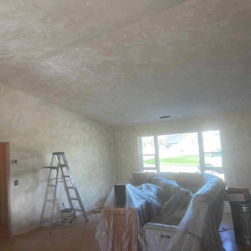 Living room interior with textured walls and ceiling, ladder, covered furniture, and a window.
