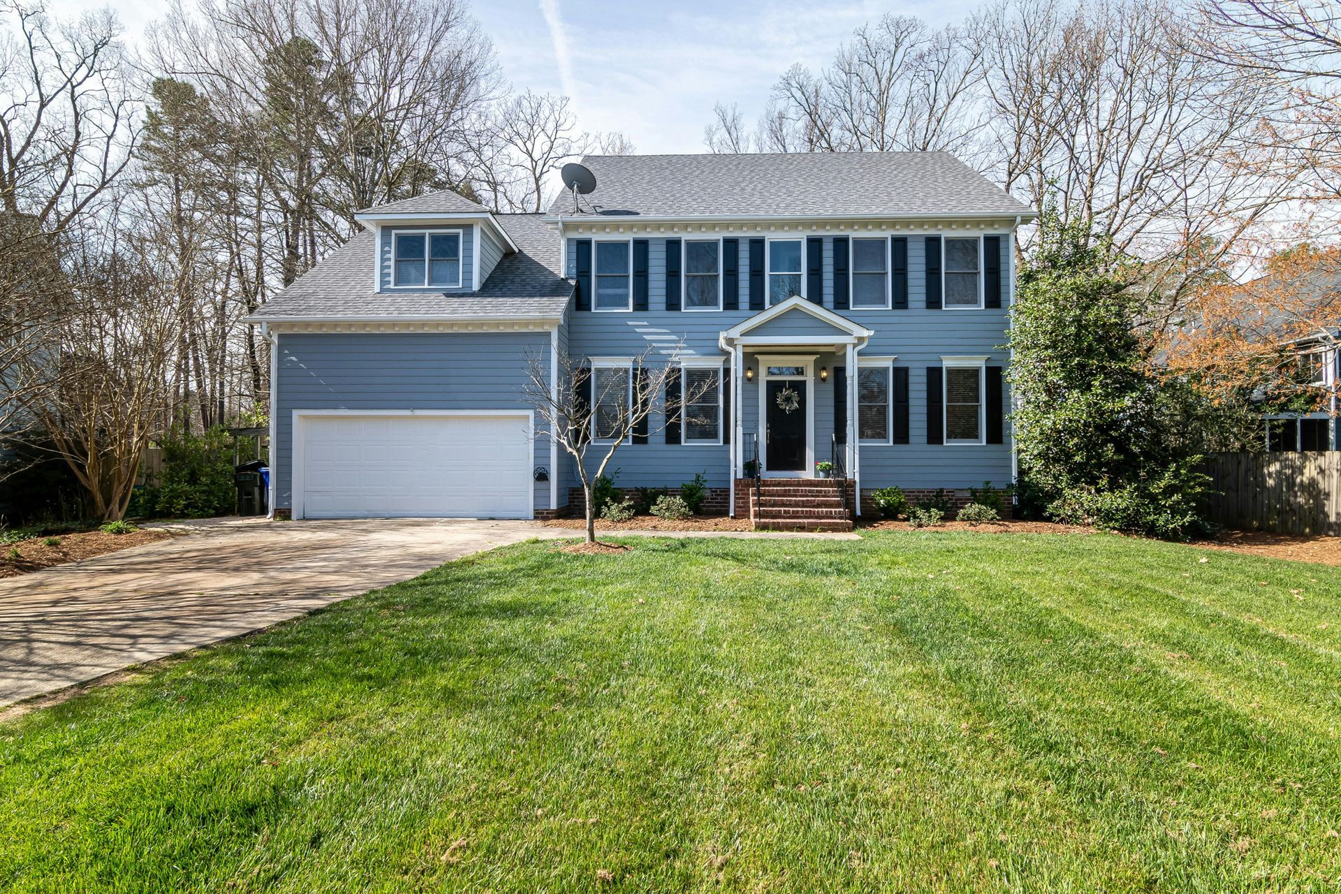 Two-story blue house with white trim, a double garage, and a front lawn in a wooded suburban setting.