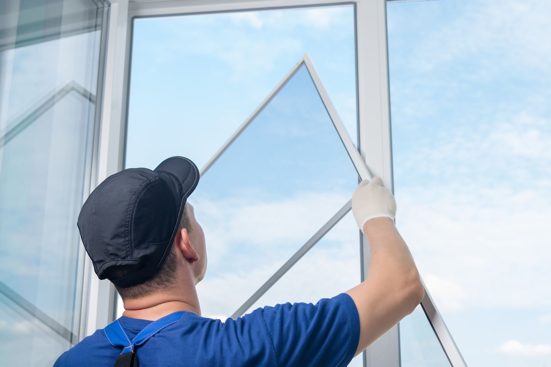 Man in blue shirt and cap installing a screen window, outdoors, against a blue sky.
