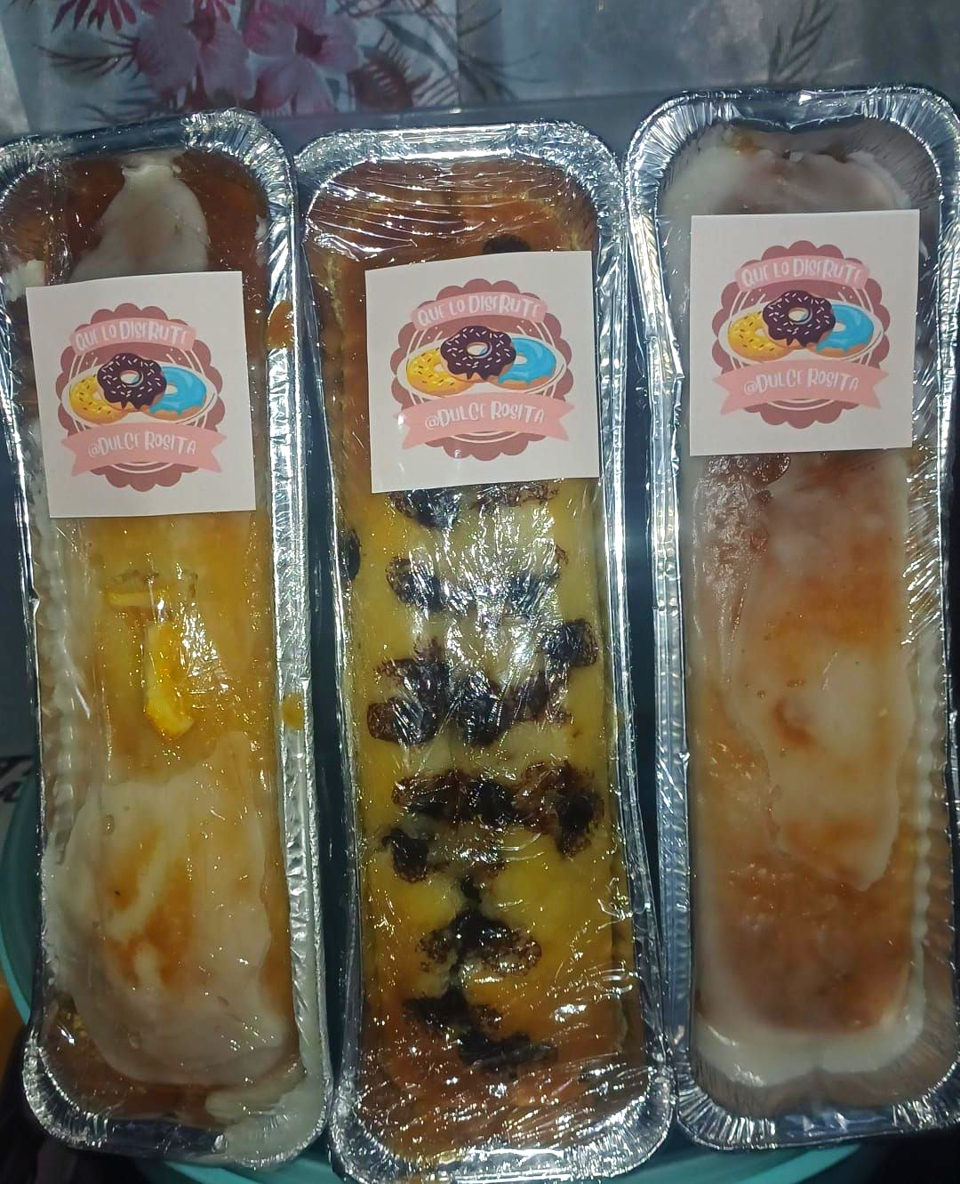 Tres bizcochos en moldes de aluminio, envueltos y con pegatinas. Amarillos, con chispas de chocolate y glaseados.