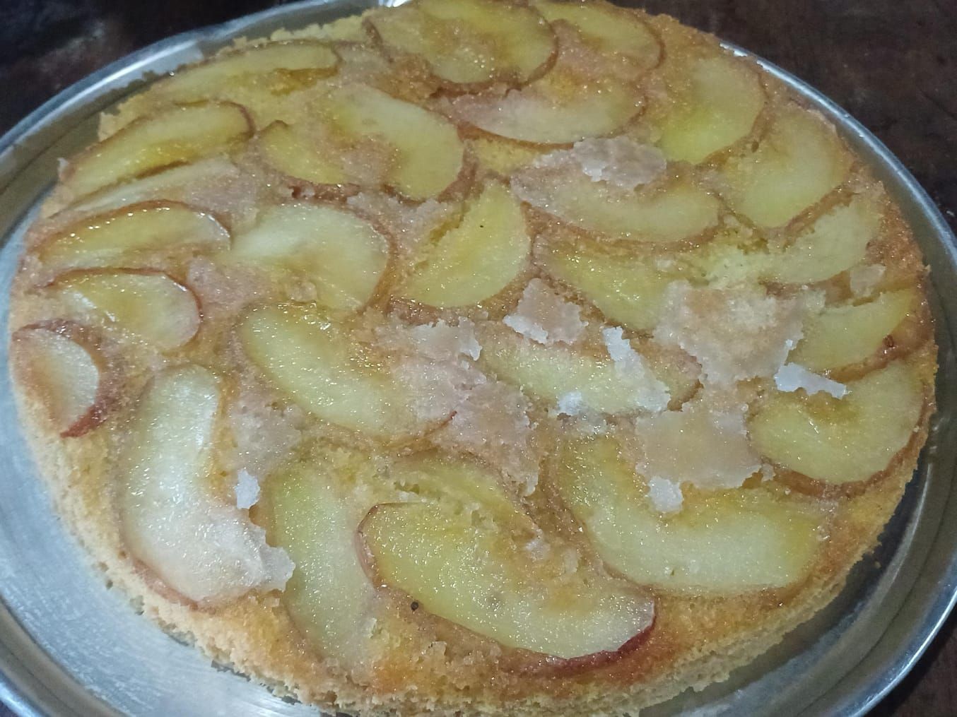Tarta de manzana, de color dorado, con rodajas de manzana dispuestas encima, en un plato redondo de metal.
