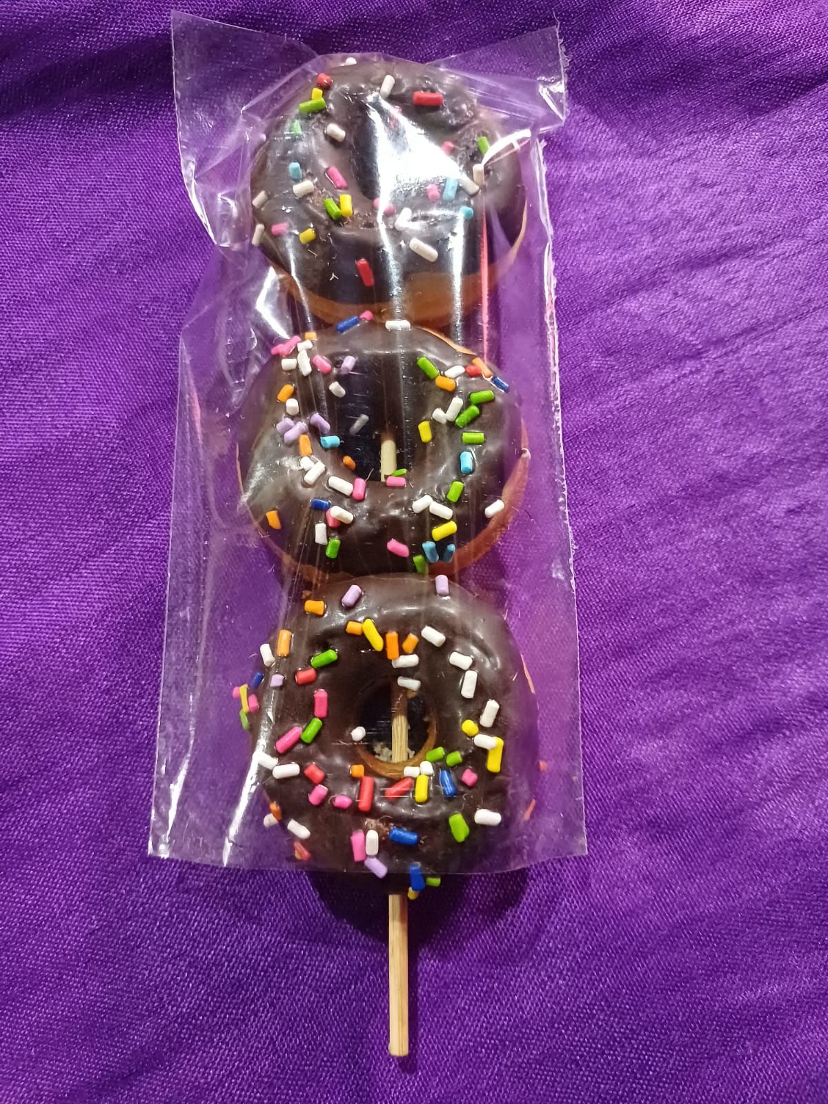 Tres donas glaseadas con chocolate y chispas en un palito, envasadas en plástico transparente, sobre tela violeta.