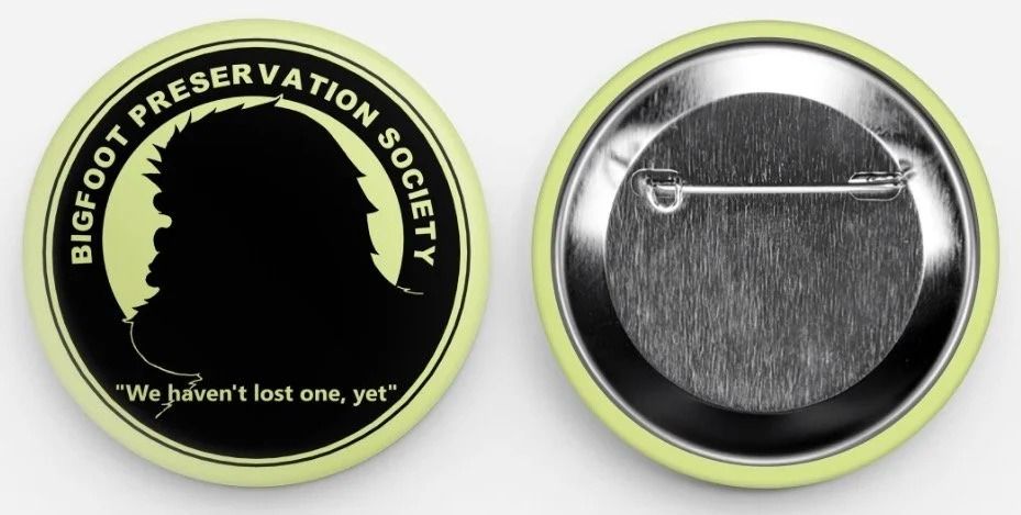 Two round buttons: one black with green rim and Bigfoot Preservation Society logo, one silver with a safety pin clasp.