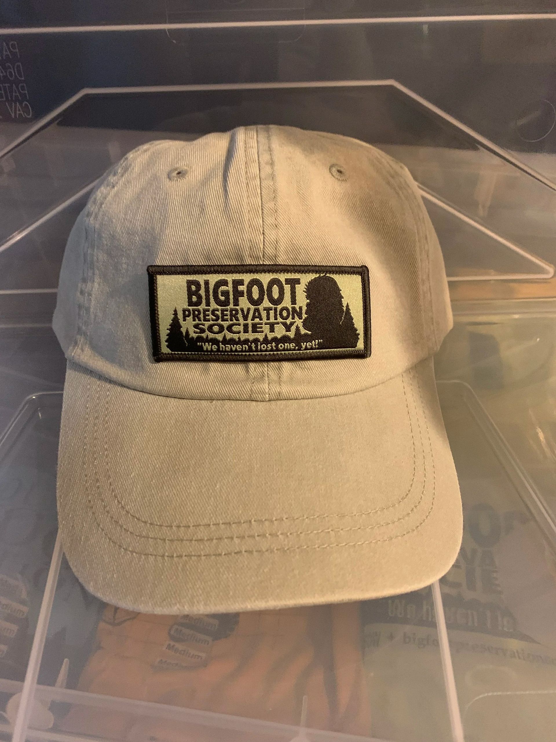 Beige Bigfoot baseball cap with a black logo patch, shown front-facing on a clear display stand.