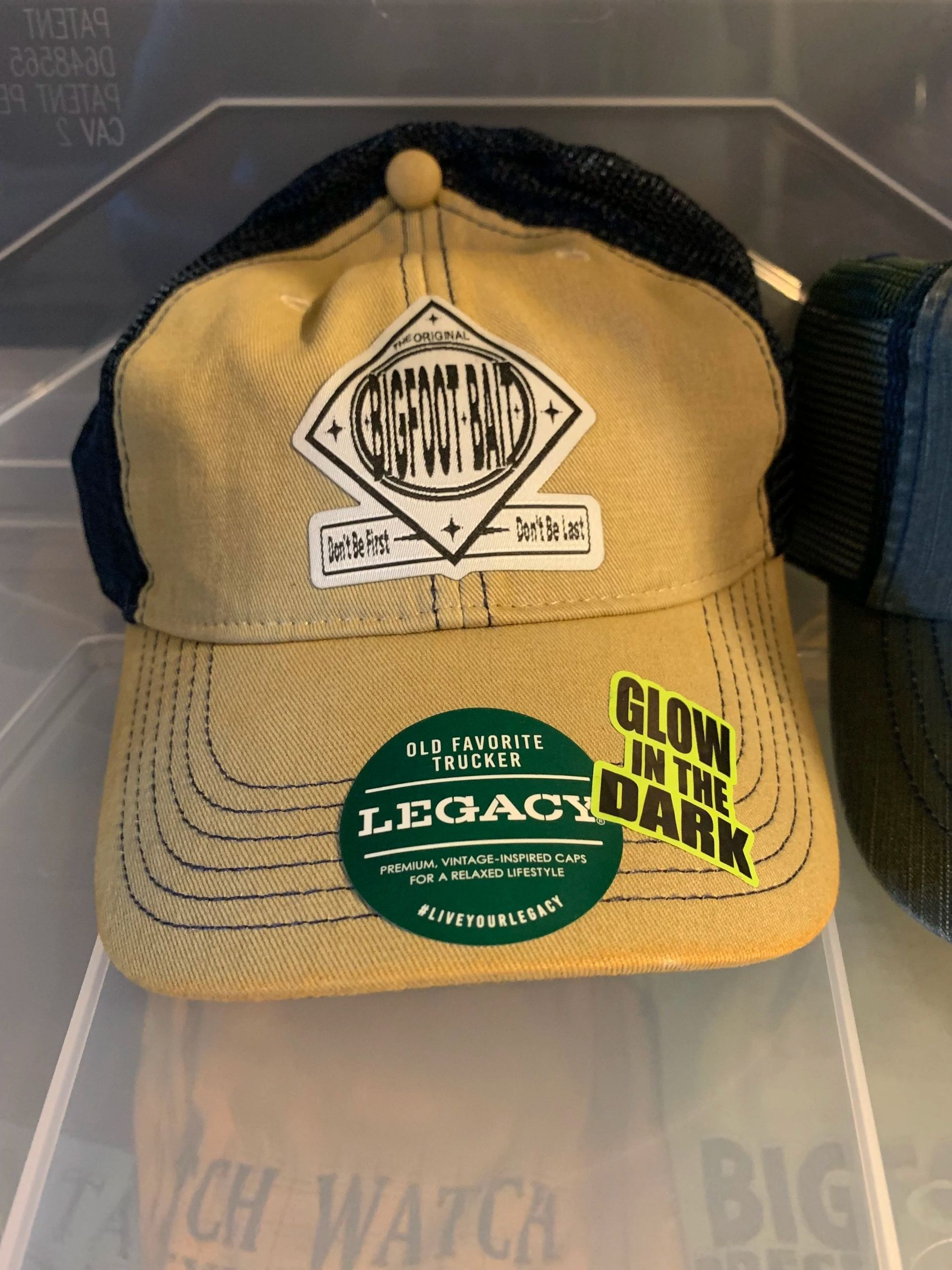 Tan trucker hat with black mesh and green stickers reading “Legacy” and “Glow in the dark”