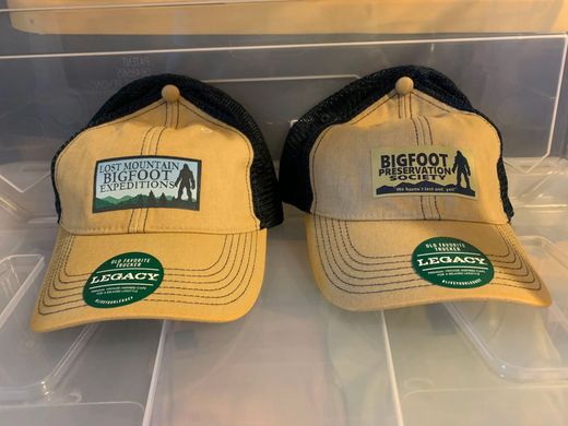 Two tan-and-black trucker hats displayed side by side in a clear plastic case