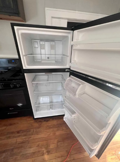 Open black refrigerator with empty shelves and freezer section. Located in a kitchen with a stove and hardwood floor.