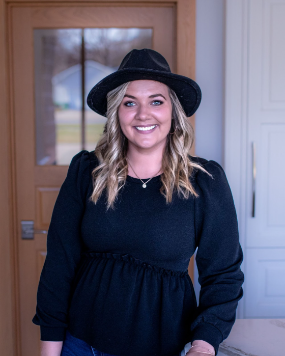 Woman wearing a black hat smiles, standing in front of a wood door. She is wearing a black top.