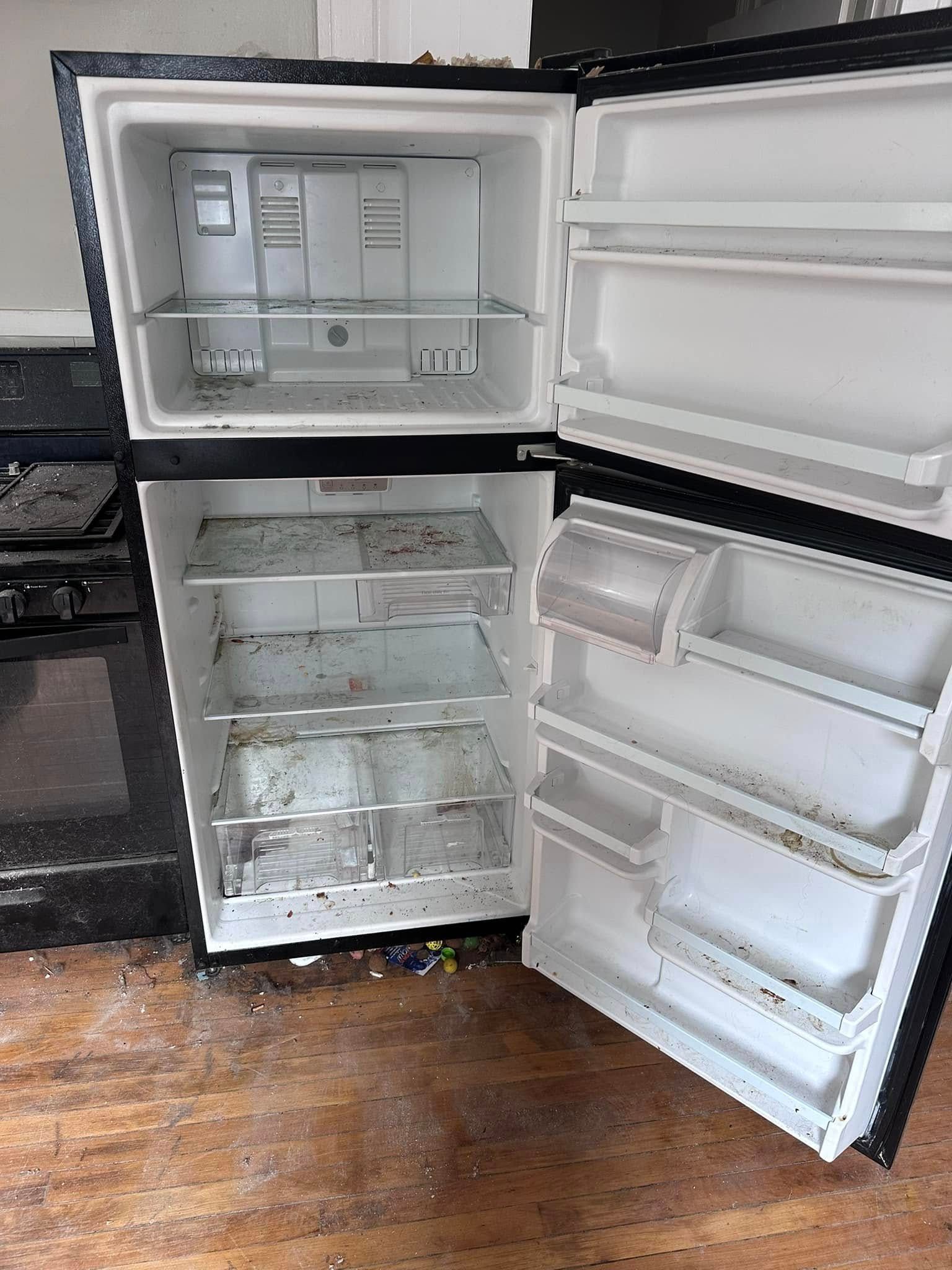 A dirty, open refrigerator with shelves and compartments, sitting on a wooden floor.
