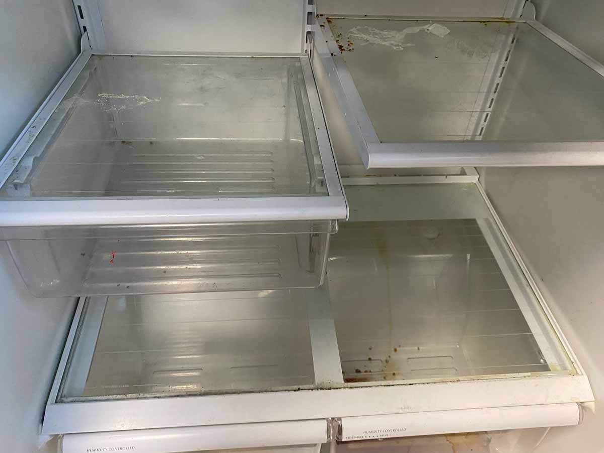 Empty refrigerator interior with glass shelves and plastic bins; some residue visible.