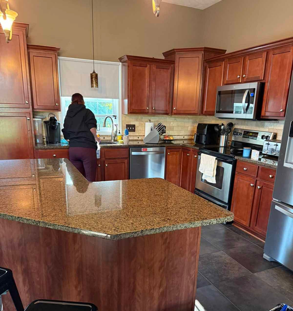 Person washing dishes in a brown-cabinet kitchen with a granite island. Stainless steel appliances.