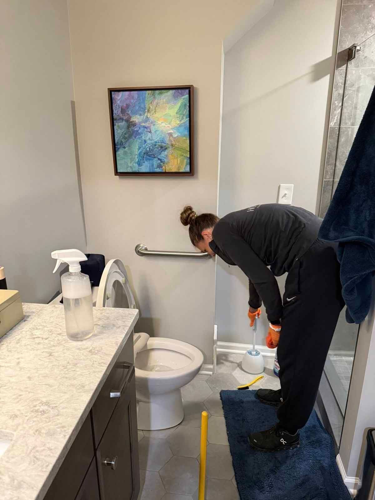 Person cleaning a bathroom toilet. They wear gloves, and dark athletic clothing.