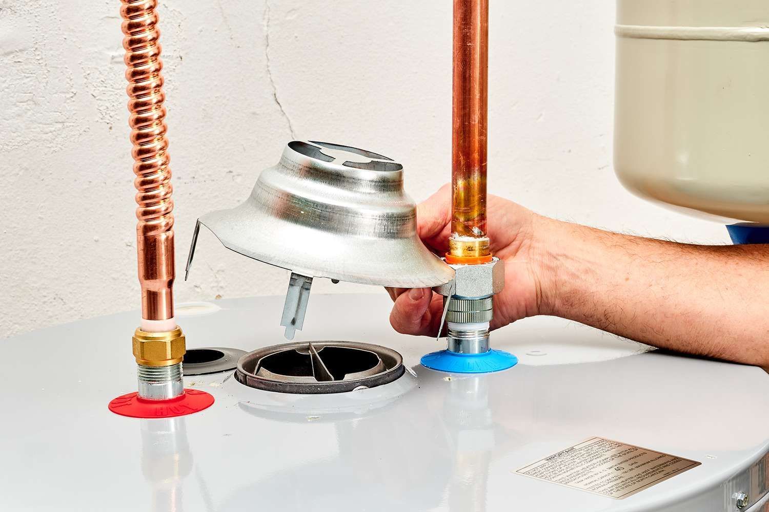 Plumber working on a water heater, holding a vent. Copper pipes, blue and red gaskets visible.