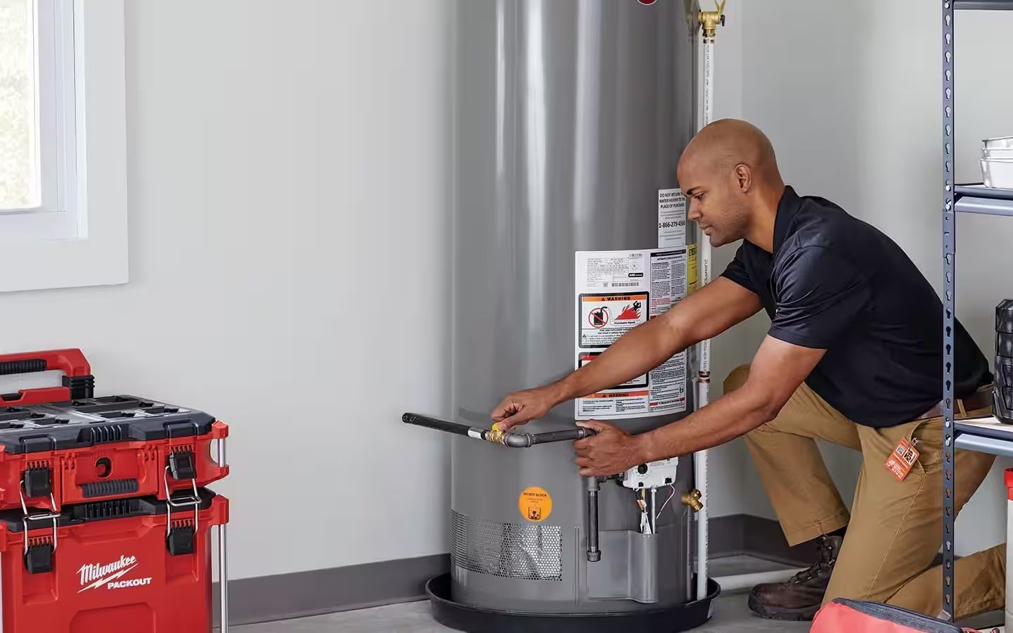 Man installing a water heater in a garage. He is kneeling, working with pipes. A toolbox is on the floor.