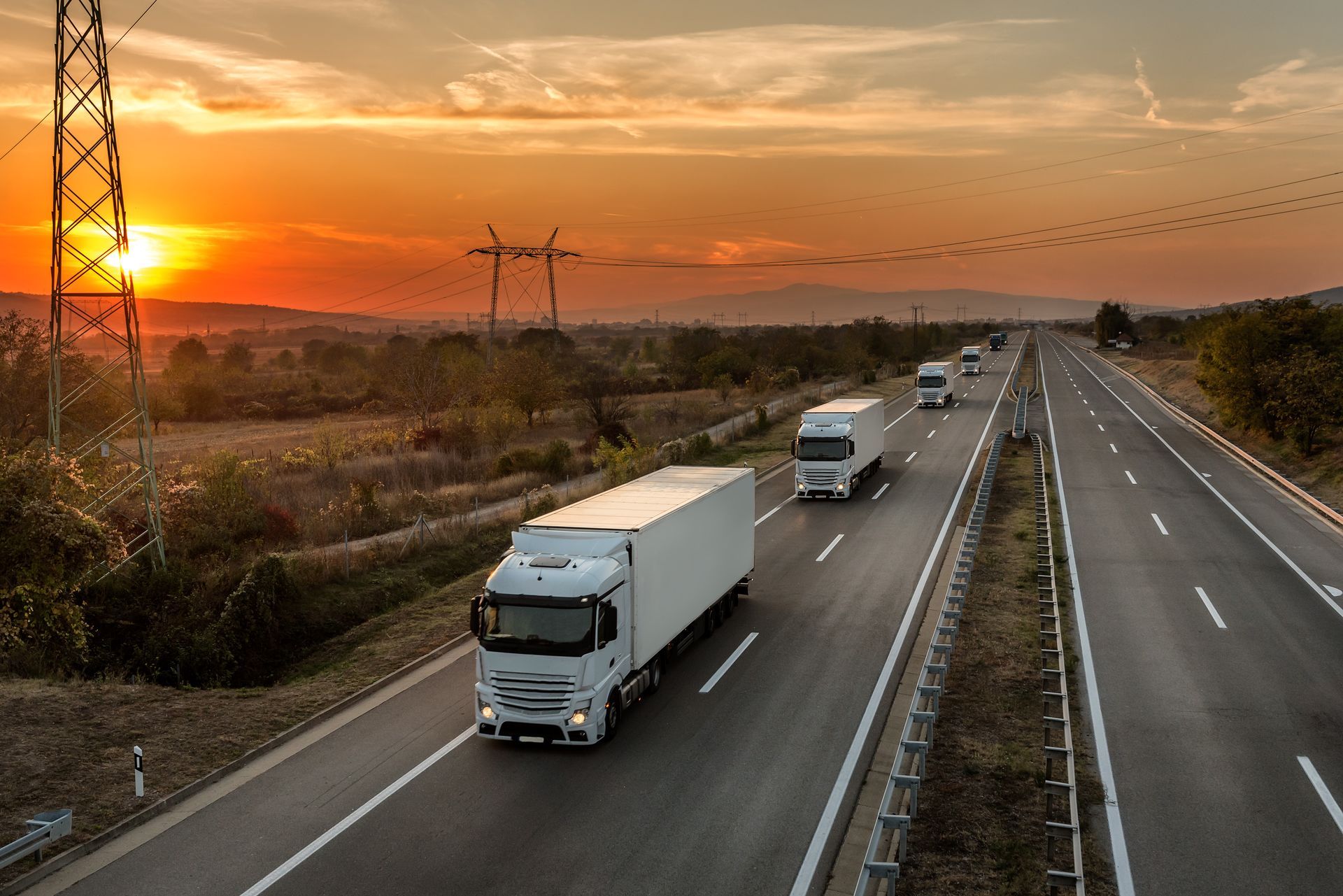 Semi-trucks and cars on a highway at sunset, with orange sky and fields beside the road.