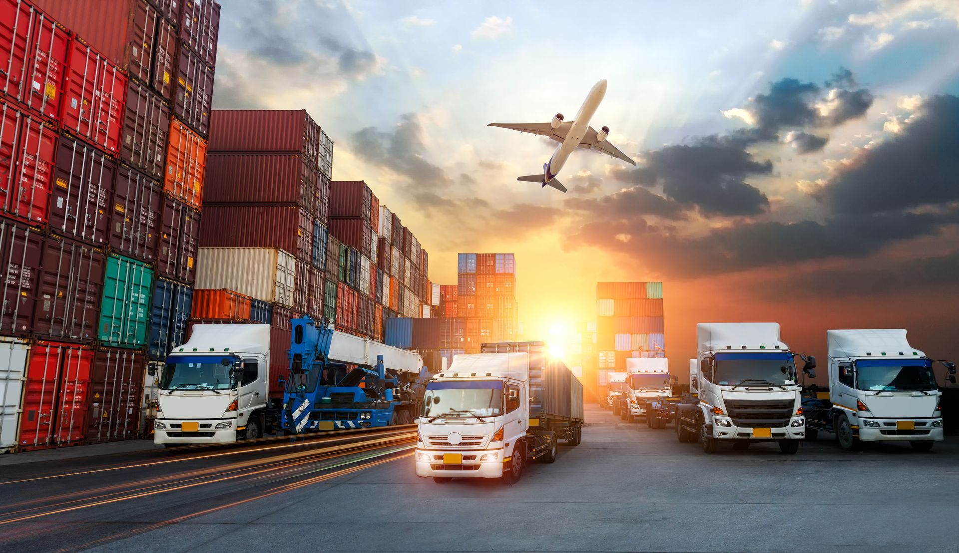 Cargo trucks at a shipping port with stacked containers and a plane overhead at sunset