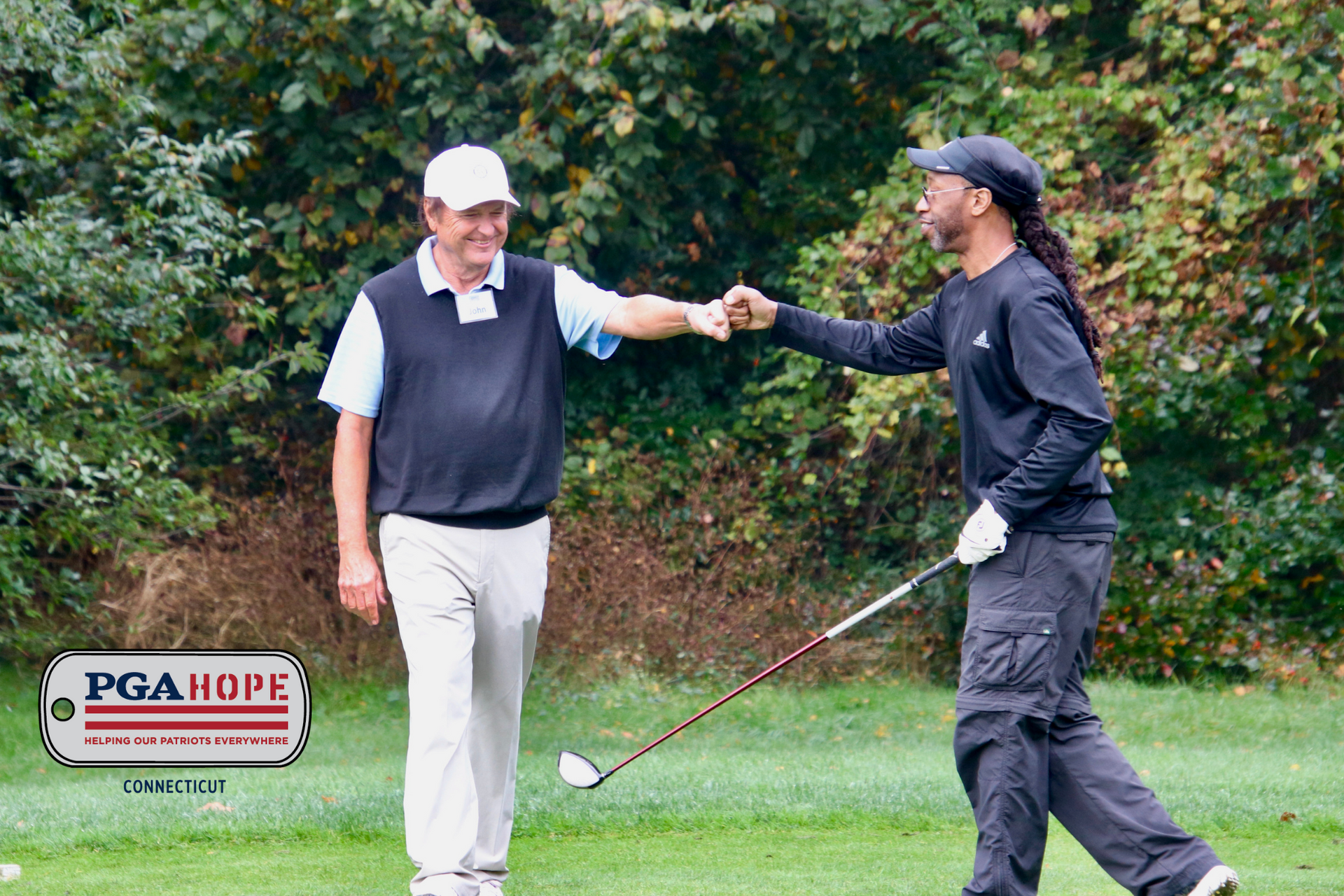 veterans fist-bump on a green fairway, one holding a club; PGA Hope sign in the lower left.